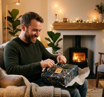 A smiling man opening a beautifully wrapped gift box in a cozy living room.
